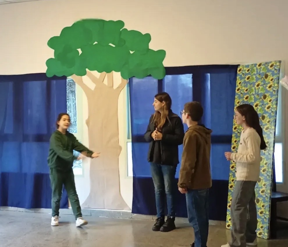 Alumnos actuando una escena frente a una escenografía con un árbol